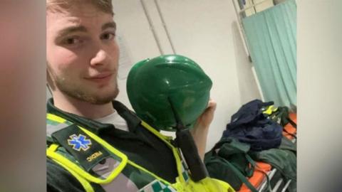 Jamie Kadolski poses in a selfie wearing an high-visibility vest. Underneath the vest is a green polo shirt and he is holding a green helmet. He is has a radio attached to the vest. In the background is a stretcher with orange mattress with bags on top.