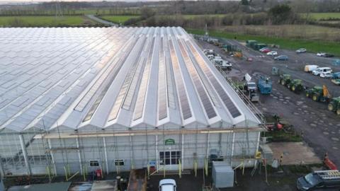 The image shows a large greenhouse with long rows of rooftop solar panels installed along its ridged roof. In front of the building is a yard area containing machinery and vehicles, including tractors and utility vehicles. Several cars are parked around the site. The surrounding landscape features open fields, hedgerows, and distant power lines.