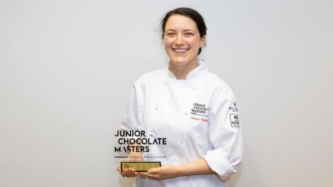 Emma Preisig wearing a white chefs apron and holding her trophy for winning the compeition. 