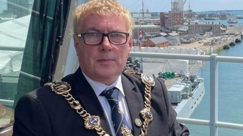 Man with blond hair and glasses facing the camera he is standing outside on a platform area with Portsmouth's harbour seen behind.