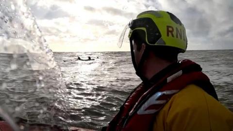 An RNLI crew member wearing yellow helmet and life jacket looking out to sea where a canoe with canoeists is in the distance