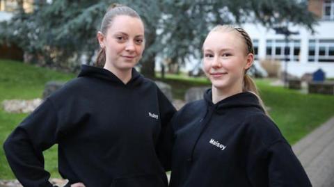Two girls with their hair in ponytails - one blonde and one brown. They are both wearing black hoodies. The girls with brown hair on the left has white writing on it that reads "Ruby" and the girl on the right has writing that reads "Maisey"