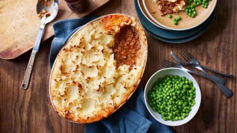 Slow cooker shepherd’s pie served with a bowl of peas on a dark brown wooden surface.