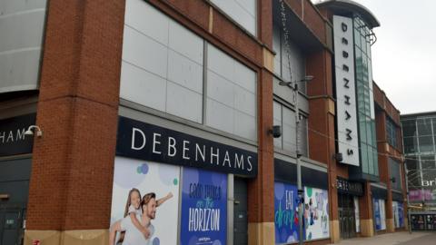 The exterior of the large Debenhams store in Sunderland. It has multiple storeys and still has adverts on the windows despite being closed since 2021. The store's name is above the windows, main entrance doors and up the middle of the building alongside a large glass frontage.