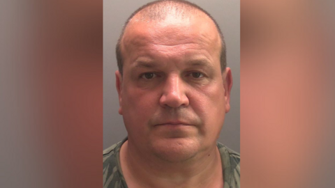 A man with short hair at back and sides and wearing a patterned green top is staring into the camera for a police custody picture.