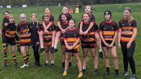 A girls' rugby team lining up on the pitch in their black, orange and red kits. Some of the girls are on the backs of other girls.