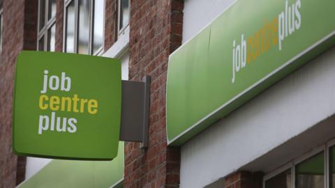 Close up of a green job centre plus sign attached to a red brick wall of an office building. The words job and plus are in lower case white lettering and centre is in yellow.