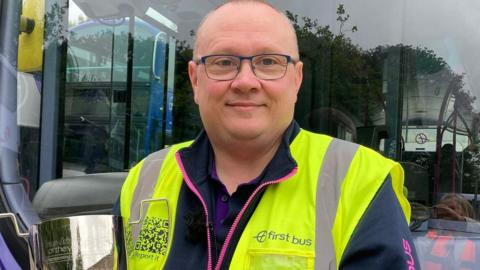 A bald man wearing a purple and blue fleece with a yellow high-vis vest over the top. He is standing in front of a bus holding a silver trophy.
