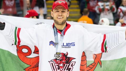 Ben Davies wearing Devils cap and jersey poses on the ice with a Wales flag