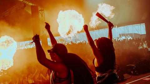 Two women on stage in a marquee raise their arms as flames shoot up on the first day of Love Saves The Day. A large crowd inside the marquee can be seen illuminated in the flames