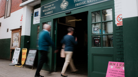 A building with a dark green entrance and a sign saying "Welcome to Wellington Market" in cream letters. A man and woman are talking through the entrance but they are blurry figures and cannot be seen in detail.