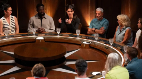 Contestants sit at a round table, with glasses of water in front of them. Most are looking towards a contestant who is talking.