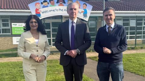 Shabina, a woman in a yellow suit, standing next to Jonathon, a man in a grey suit and Andrew Pakes, a man in a blue jumper and jeans, smiling for camera. 