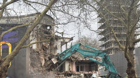A green crane tears into a half-destroyed large building. Two trees are in the foreground and a tower with scaffolding is in the distance.
