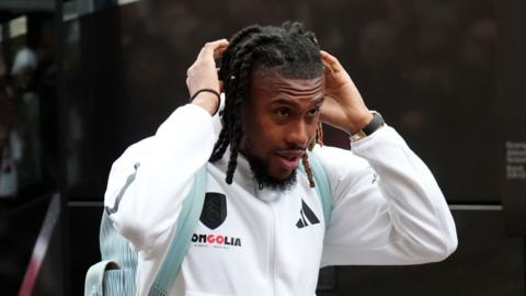 A man with braids wears a white tracksuit. His hands are raised to his hair