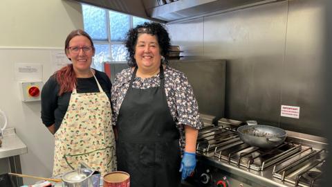 Ioni Drake and Mandy Harding (left). Iona is wearing a black top and light yellow apron. Mandy is wearing a floral top and black apron and blue gloves. They are stood in a kitchen.