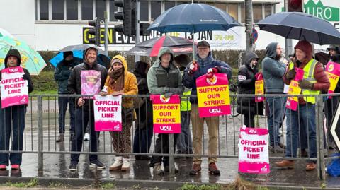 The picket line which has 12 teachers standing behind a meatl gate holding signs which read: OFFICIAL PICKET LINE and SUPPORT OUR STRIKE and they are branded by UCU. It is raining so the teachers are wearing anoraks and have umbrellas. Behind them is a Truro and Penwith College building.
