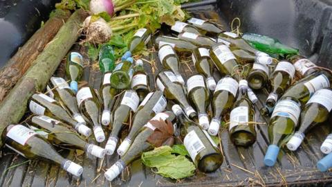 The bed of a pickup truck filled with empty glass wine bottles. The pile of green bottles lies next to two wooden logs and large vegetables.