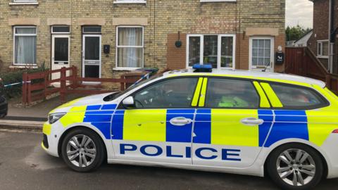 A police car parked outside light brick-built terraced houses, with police tape visible on a brown fence.