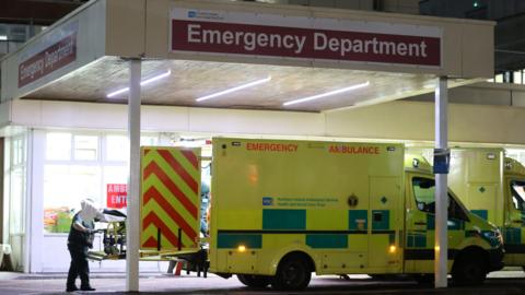 Ambulance parked under a covered porch, with Emergency Department written across the top. A paramedic is taking a trolley out of the ambulance