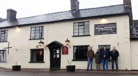 A white building with black framed windows and door, a black tiled roof and four people standing outside. There are two vintage lamps on the wall on each side of the entrance door. There is also a burgundy sign with Crown Inn written on it. Above the four people there is a black banner attached to the wall, saying "Newcastle's Crown Inn - Reclaiming the Crown Inn for the Community".