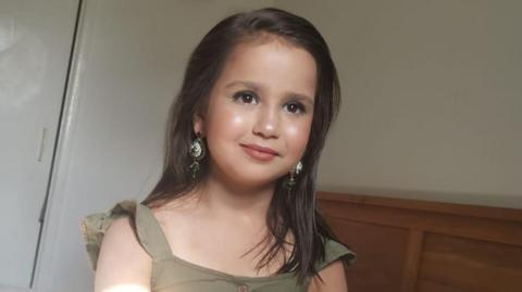 Sara Sharif staring off camera dressed in a light coloured dress and wearing earrings