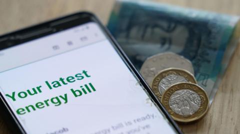 An energy bill statement on a mobile phone screen. A folded £5 note and some coins are alongside the phone.
