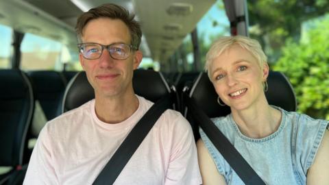 Two people sit beside each other on a coach, both wearing seatbelts and facing the camera. The man on the left wears glasses and a light pink T-shirt while the woman on the right has short blonde hair and wears a sleeveless denim top. The coach interior is visible with rows of dark-coloured seats behind them and sunlight streams through large windows, with greenery outside.
