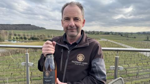 A man with grey hair is wearing a brown and grey jacker with a circular gold logo on the breast. white writing on it says "halfpenny green wine estate". He is holding a bottle of rosé wine. He is on a balcony and beyond him is a vineyard with green grass and vines growing