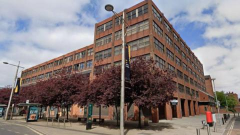 A Google Streetview screenshot of Vancouver House in Middlesbrough. The large six-storey office block  is built in red brick with large rectangular windows on each floor. The building gives onto a main road where there is a bus stop.