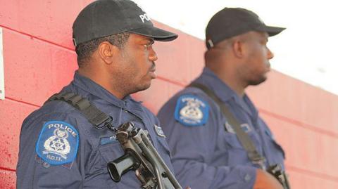 Two armed policemen in Port of Spain, Trinidad, seen in profile wearing black baseball caps and blue shirts.