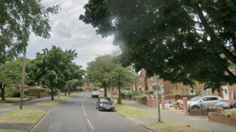 A suburban road lined with trees and houses. Parked cars, lamp-posts and other street furniture can be seen.