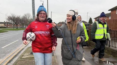 A woman wearing a blue Hull KR bobble hat and a red Hull KR jersey is walking towards the camera with a rugby ball under her arm. She is wearing glasses with red frames and has red hair, she's being interviewed by a blonde woman who is holding a phone with BBC Radio Humberside branding. They are flanked by other walkers wearing hi-vis vests as they walk down a residential street. 