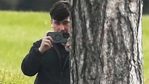 A man in a dark jacket and blue jeans stands partially concealed by a tree trunk and holds a phone up in two hands as if filming and has a white earphone in his right ear