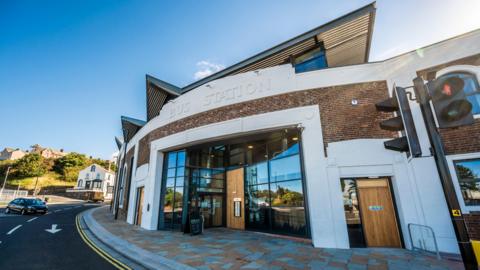 The outside of the Bus Station building. It has curved frontage with glass windows and wooden door. The words "Bus Station" are above the entrance.