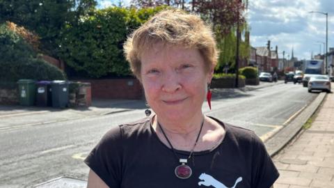 A woman with a red pendant necklace, a black Puma t-shirt, two earrings, her left one is a skull, her right is a chilli. She has light brown short hair. In the background is a road, with cars, and houses in the distance, to the right
