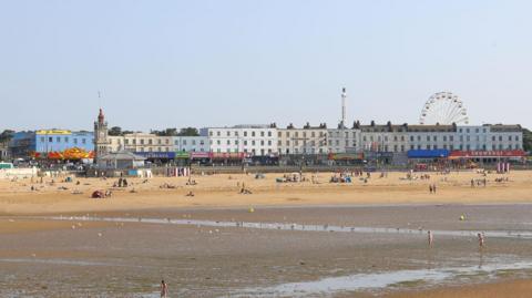 A picturesque English seaside town viewed from out at sea. Visible are golden sands, a row of houses and attractions. 