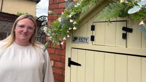 Natalie Brook from Rhodesia is stood in front of her cake shed.
She has blond hair and is wearing glasses. 