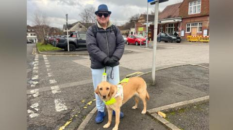 Paul Goddard, a blind railway campaigner from East Sussex, pictured at Crowborough train station with his golden coloured guide dog. Paul is wearing a blue fedora, blue trainers, puffer jacket and pale blue jeans. He is also wearing black gloves and glasses.