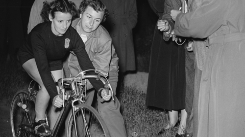 A black and white photo of Eileen Sheridan setting off on a record-breaking run from London to Oxford. She is wearing a dark top and shorts, as crowds stand around her