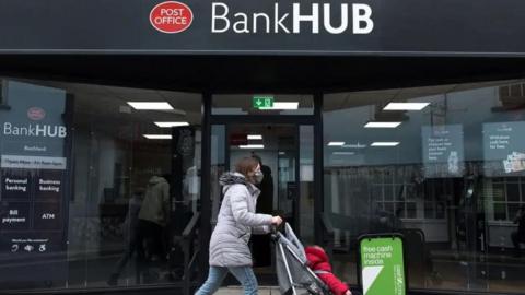 A woman wearing a mask is pushing a child in a buggy past a banking hub which also has the red Post Office sign. 