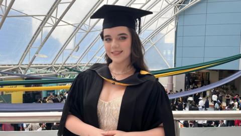 A woman with long dark hair is wearing a graduation gown and cap smiling