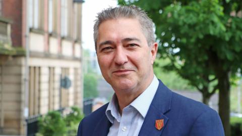 Mark Wynn with short grey hair smiling wearing a blue blazer, checked blue and white shirt and red badge outside Lancashire County Council's County Hall in Preston with trees in the background 