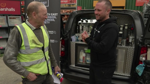 James Fildes, in black hoody and trousers is smiling and talking to a man in khaki jumper, high visibility vest and jeans. They are standing in front of the van in Durham, which is equipped with a coffee machine, fridge with milk and cream and disposable paper cups. 