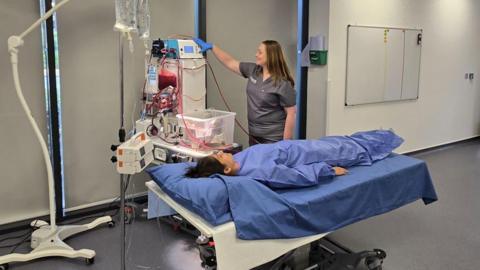 A woman wearing blue scrubs and lying on a hospital bed. A woman in grey scrubs is standing next to her, with her hand on a machine.