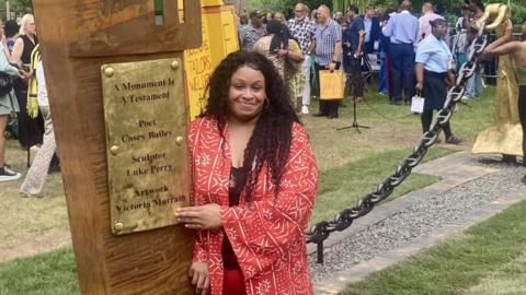 A young black woman with dark curly hair points at the gold plated sign on the Windrush monument 'A Monument is a Testament', which shows her name. She is wearing a red kimono style top with a white pattern on it