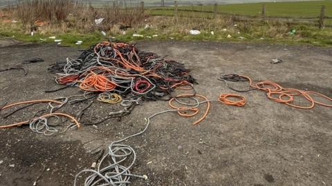 A large amount of cables and cable sleeving coloured orange, grey, black and red have been dumped on a country road. There is other waste that has been dumped in the background. 