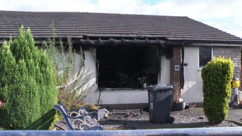 A white stone bungalow has a black, sooty hole where a window used to be. Soot marks can also be seen to have reached the roof.