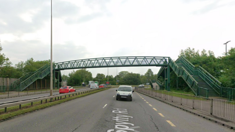 Spytty Road, a multi-lane road runs beneath a green metal pedestrian footbridge with staircases on both sides. Several cars and a white van travel along the road, and trees line both sides.