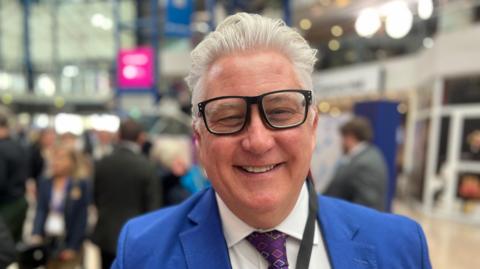 Kevin Bentley with grey hair and large black-framed glasses smiling at the camera in a bright blue suit. He is in a conference hall which is out of focus behind him.